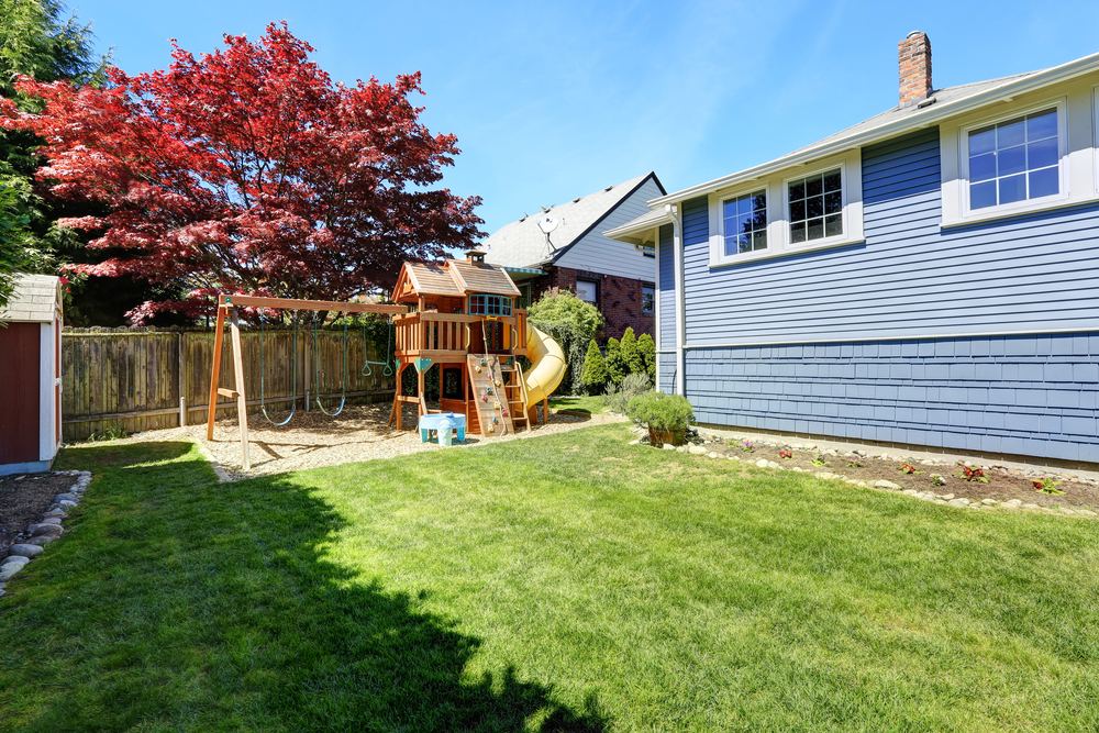 small blue bungalow fenced back yard with swing set attached to treehouse. lawn is lush and green and sky is blue.